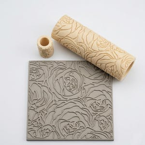 May include: A beige cylindrical clay roller and a smaller matching cylindrical piece, both featuring a deeply carved rose pattern, positioned next to a square gray clay slab embossed with the same intricate floral design across its surface.