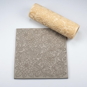 May include: A cylindrical beige rolling pin with a deeply carved floral and hummingbird pattern, positioned above a square slab of gray clay that displays the same intricate embossed design of blooming flowers and hummingbirds in flight.