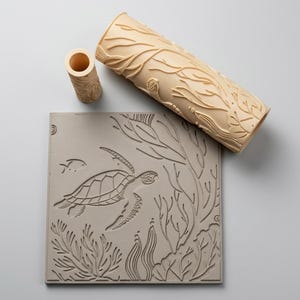 May include: A light beige cylindrical clay roller and a smaller matching tube, both featuring an embossed sea turtle and seaweed pattern, positioned above a square gray clay slab with the same detailed marine life design pressed into its surface.