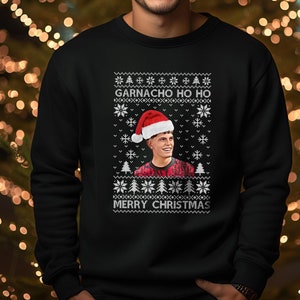 Könnte beinhalten: Schwarzes Sweatshirt mit weihnachtlichem Design. Das Design enthält den Text "GARNACHO HO HO" und "MERRY CHRISTMAS" in Weiß, zusammen mit einem Porträt einer Person mit Weihnachtsmütze. Der Hintergrund ist ein Bokeh aus Weihnachtslichtern.