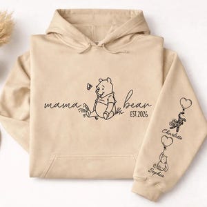 May include: Beige hoodie with a hood and drawstring. The front features a Winnie the Pooh graphic with the words "mama bear" and "est. 2026". The right sleeve has Winnie the Pooh characters holding balloons with names.