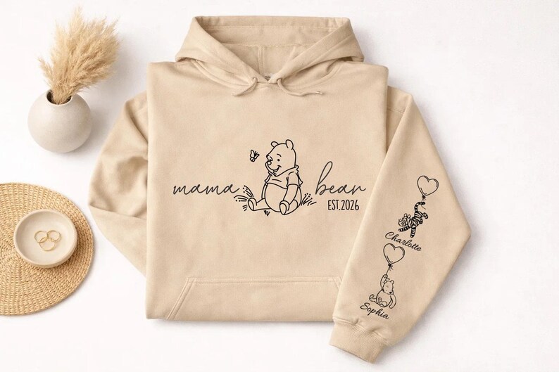 May include: Beige hoodie with a hood and drawstring. The front features a Winnie the Pooh graphic with the words "mama bear" and "est. 2026". The right sleeve has Winnie the Pooh characters holding balloons with names.