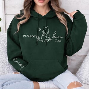 May include: Dark green hoodie with a white graphic of Winnie the Pooh and the words "mama bear" and "EST 2026". The hoodie has a front pocket and a drawstring hood. A small graphic and the name "Evelyn" are on the left sleeve.