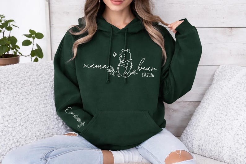 May include: Dark green hoodie with a white graphic of Winnie the Pooh and the words "mama bear" and "EST 2026". The hoodie has a front pocket and a drawstring hood. A small graphic and the name "Evelyn" are on the left sleeve.