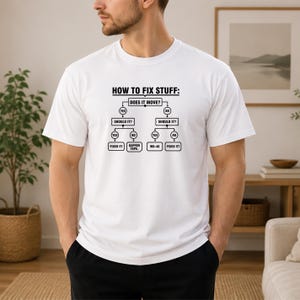 May include: A white short-sleeved t-shirt featuring a black graphic print of a flowchart titled HOW TO FIX STUFF. The diagram includes decision boxes labelled DOES IT MOVE, SHOULD IT, FIXED IT, GAFFER TAPE, and WD-40 with connecting arrows.