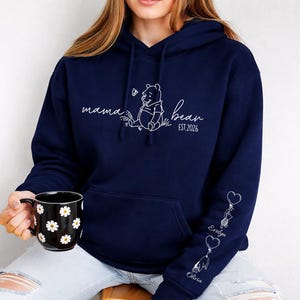 May include: Navy blue hoodie with a white graphic of Winnie the Pooh and the words "mama bear" and "est. 2026". The sleeve features heart-shaped balloons with names. A black mug with white daisy patterns is held in the hand.