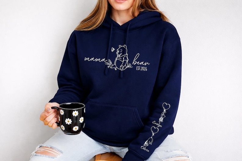 May include: Navy blue hoodie with a white graphic of Winnie the Pooh and the words "mama bear" and "est. 2026". The sleeve features heart-shaped balloons with names. A black mug with white daisy patterns is held in the hand.
