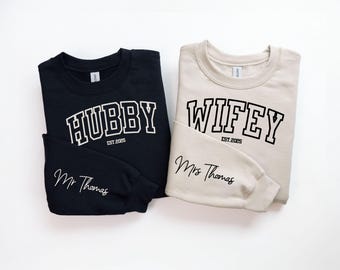 May include: Two sweatshirts, one black and one beige, with the words "Hubby" and "Wifey" in a collegiate font. The text "Est. 2025" appears below each word. The black sweatshirt has the text "Mr. Thomas" on the sleeve, and the beige sweatshirt has the text "Mrs. Thomas" on the sleeve.