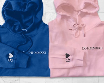 May include: Two hooded sweatshirts, one blue and one pink, with a heart and roman numeral date embroidered on each sleeve. The blue sweatshirt has a white heart and the date "I-II-MMXXI" on the left sleeve. The pink sweatshirt has a black heart and the date "IX-I-MMXXII" on the right sleeve.
