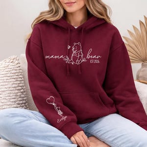 May include: A maroon hoodie with a kangaroo pocket and a drawstring hood. The front features white script that says "mama bean" with a Winnie the Pooh graphic and "EST. 2026". The sleeve has a graphic of a cat holding a heart balloon and the name "Evelyn".