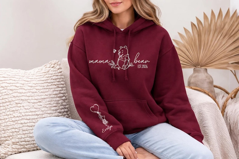 May include: A maroon hoodie with a kangaroo pocket and a drawstring hood. The front features white script that says "mama bean" with a Winnie the Pooh graphic and "EST. 2026". The sleeve has a graphic of a cat holding a heart balloon and the name "Evelyn".
