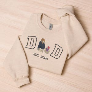 May include: A beige sweatshirt with the word "DAD" in black block letters, with an illustration of a father and child. The text "EST. 2024" is below the illustration. The sweatshirt is folded on a light wood surface.