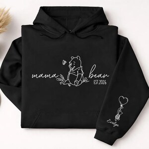 May include: Black hoodie with white line art. The front features the words "mama bear" with a Winnie the Pooh illustration and "bear est. 2026". The sleeve has a cat holding a heart balloon and the name "Evelyn".