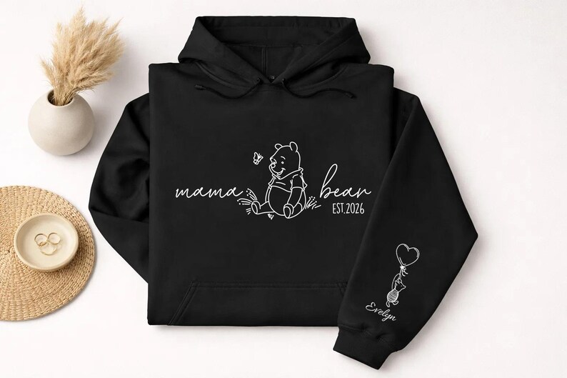 May include: Black hoodie with white line art. The front features the words "mama bear" with a Winnie the Pooh illustration and "bear est. 2026". The sleeve has a cat holding a heart balloon and the name "Evelyn".