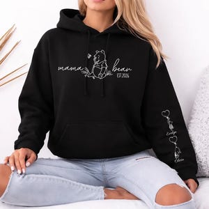 May include: Black hoodie with a white graphic of Winnie the Pooh and the words "mama bear" and "est. 2026". The sleeve features heart-shaped keychains with names. A comfortable, casual sweatshirt.