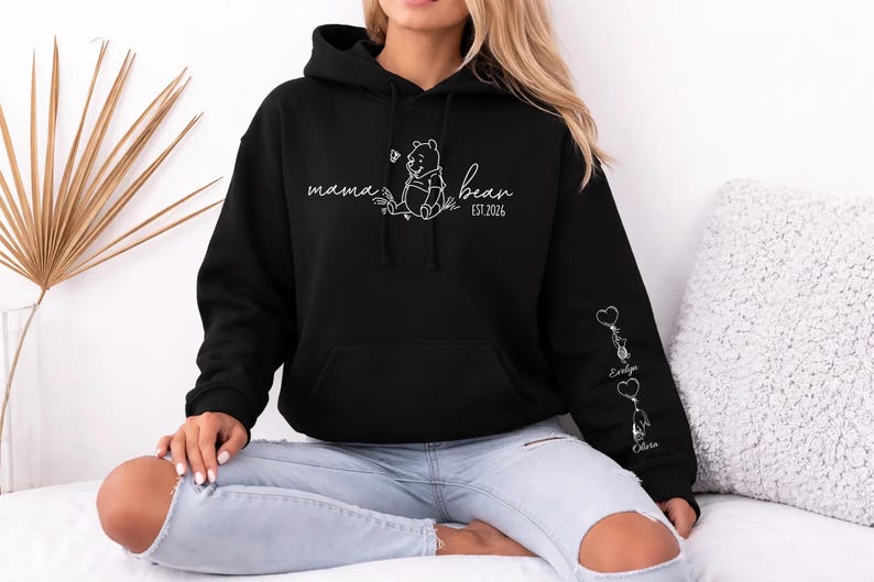 May include: Black hoodie with a white graphic of Winnie the Pooh and the words "mama bear" and "est. 2026". The sleeve features heart-shaped keychains with names. A comfortable, casual sweatshirt.