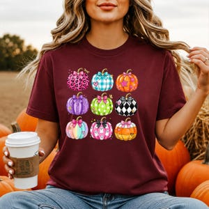May include: A maroon short-sleeve t-shirt featuring a graphic print of nine stylised pumpkins arranged in a grid. Each pumpkin displays a unique pattern, including leopard print, plaid, polka dots, and ghosts, all accented with colourful bows.