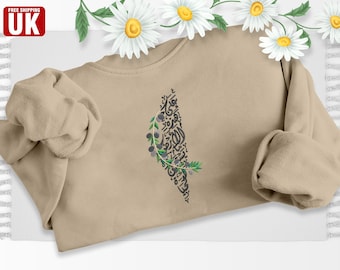 May include: A beige sweatshirt with an olive branch design in the shape of Palestine. The design is made up of Arabic calligraphy and olive branches with black olives.