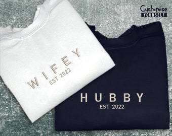 May include: Two sweatshirts, one white and one navy blue, with the words "WIFEY EST 2022" and "HUBBY EST 2022" in gold lettering.