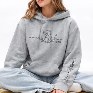 May include: A light gray hoodie with a front pocket and a drawstring hood. The front features a Winnie the Pooh graphic with the words "mama bear" and "EST 2026". The sleeve has three heart-shaped balloons with names.