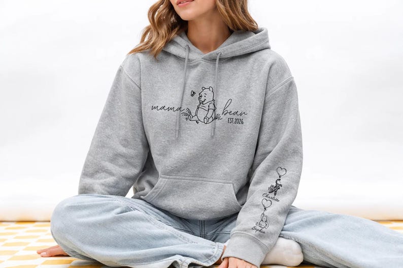 May include: A light gray hoodie with a front pocket and a drawstring hood. The front features a Winnie the Pooh graphic with the words "mama bear" and "EST 2026". The sleeve has three heart-shaped balloons with names.