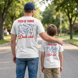 Pode incluir: Duas camisetas brancas com designs correspondentes. A camisa do adulto diz "Fixer of Things" com ferramentas e "Dads club". A camisa da criança diz "I Break Stuff" com brinquedos e "Kids club".