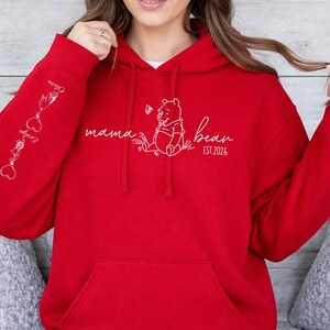 May include: A red hoodie with a kangaroo pocket and a drawstring hood. The front features a white graphic of Winnie the Pooh with the text "mama bear EST. 2026". The left sleeve has white graphics and names. A cozy and casual apparel item.
