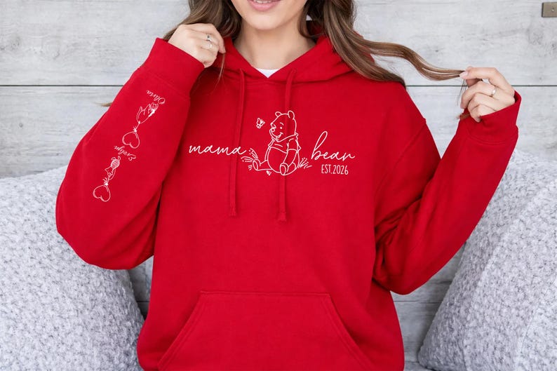 May include: A red hoodie with a kangaroo pocket and a drawstring hood. The front features a white graphic of Winnie the Pooh with the text "mama bear EST. 2026". The left sleeve has white graphics and names. A cozy and casual apparel item.