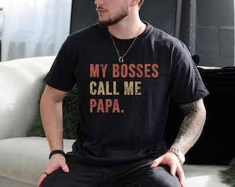 May include: Black t-shirt with the text "MY BOSSES CALL ME PAPA" in red and yellow lettering. The shirt is worn by a person with a beard and a gold necklace.