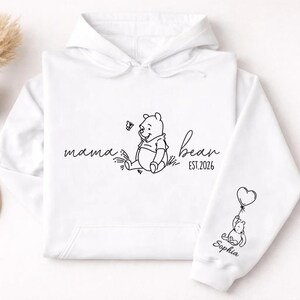 May include: White hoodie with a black line drawing of a bear, a butterfly, and the words "mama bean EST.2026". The sleeve features a bear holding a heart-shaped balloon and the name "Sophia".