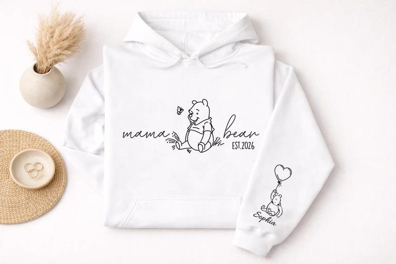 May include: White hoodie with a black line drawing of a bear, a butterfly, and the words "mama bean EST.2026". The sleeve features a bear holding a heart-shaped balloon and the name "Sophia".