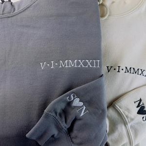 May include: Two gray and beige hooded sweatshirts with embroidered text. The gray sweatshirt has the text "V.I.MMXXII" and a heart with the letter "S" on the sleeve. The beige sweatshirt has the text "V.I.MMXXII" and a heart with the letter "N" on the sleeve.