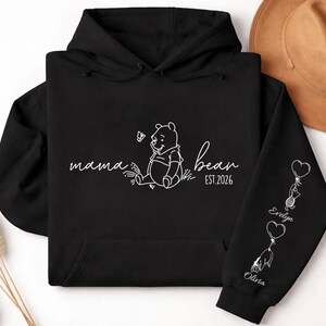 May include: Black hoodie with white script reading "mama bear" and a Winnie the Pooh graphic. The right sleeve features heart-shaped balloons with names. A brown hat is visible in the background.