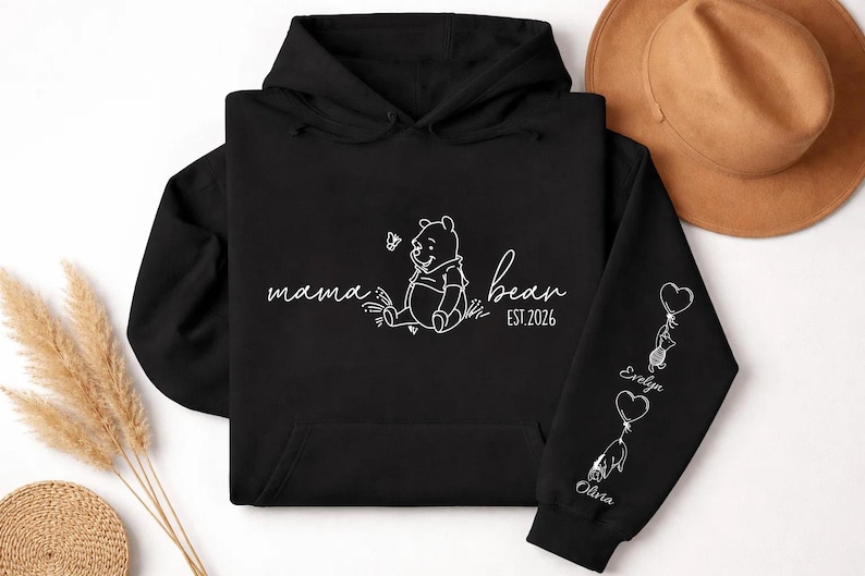 May include: Black hoodie with white script reading "mama bear" and a Winnie the Pooh graphic. The right sleeve features heart-shaped balloons with names. A brown hat is visible in the background.