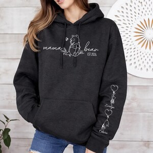 May include: Dark gray hoodie with a front pocket and a hood. The front features the words "mama bear" with a Winnie the Pooh graphic and the year "EST. 2026". The left sleeve has heart-shaped balloons with names.