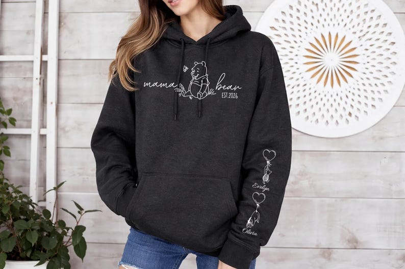 May include: Dark gray hoodie with a front pocket and a hood. The front features the words "mama bear" with a Winnie the Pooh graphic and the year "EST. 2026". The left sleeve has heart-shaped balloons with names.