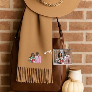 May include: A beige scarf with a fringed edge and an embroidered couple design. A photo of the couple is next to the scarf. A beige hat with a gold chain and a brown leather bag are also in the image.