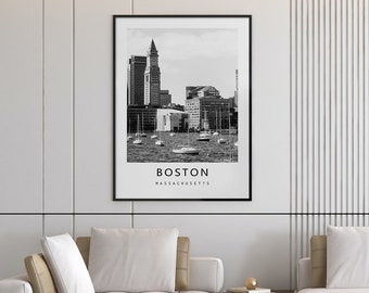 May include: A black and white framed print of the Boston skyline hangs above a modern sectional sofa. The print features the city's architecture and the text "BOSTON MASSACHUSETTS". The living room has a neutral color palette with a wooden coffee table and decorative accents.