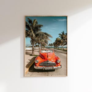 May include: Framed print featuring a vintage red convertible parked on a road lined with palm trees. The car has a chrome grill and headlights. The sky is blue with a few clouds. The print is in a wooden frame.