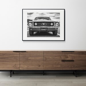 Pode incluir: Impress&atilde;o emoldurada em preto e branco de um Ford Mustang cl&aacute;ssico. A frente do carro est&aacute; centrada, mostrando far&oacute;is, grade e placa. A impress&atilde;o &eacute; exibida acima de um arm&aacute;rio de madeira com pernas e puxadores pretos.