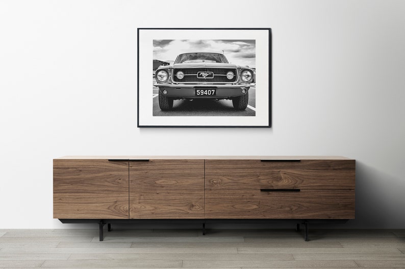 Pode incluir: Impress&atilde;o emoldurada em preto e branco de um Ford Mustang cl&aacute;ssico. A frente do carro est&aacute; centrada, mostrando far&oacute;is, grade e placa. A impress&atilde;o &eacute; exibida acima de um arm&aacute;rio de madeira com pernas e puxadores pretos.