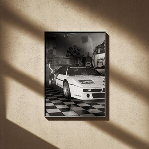 May include: Black and white framed print of a vintage race car in a garage setting. The car is white with black accents and sponsor logos, parked on a checkered floor. The print is displayed on a beige wall with shadows.