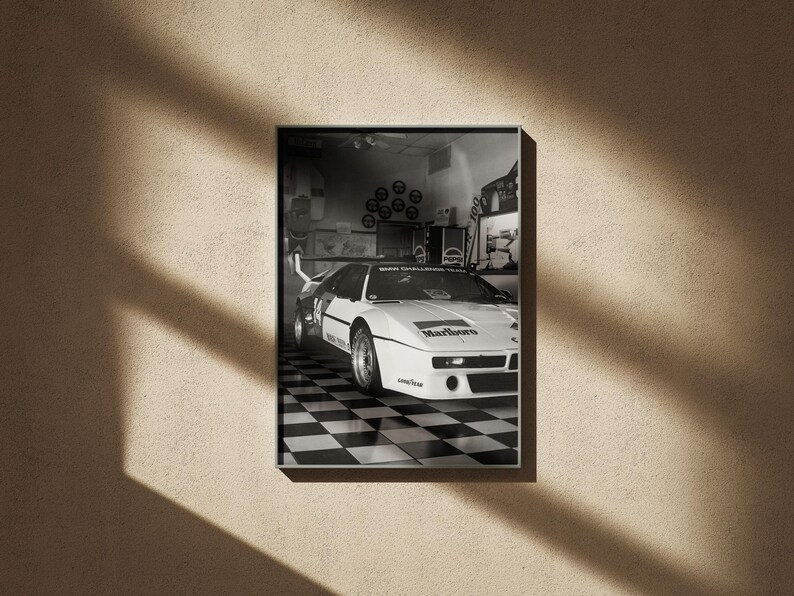 May include: Black and white framed print of a vintage race car in a garage setting. The car is white with black accents and sponsor logos, parked on a checkered floor. The print is displayed on a beige wall with shadows.