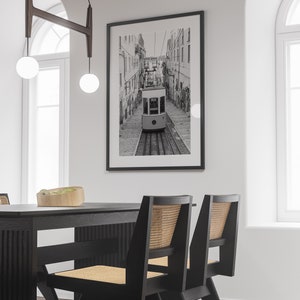 May include: Black and white framed print of a Lisbon tram traveling down a cobblestone street. The image is displayed on a white wall above a black dining table with matching chairs. The chairs have woven cane backs.