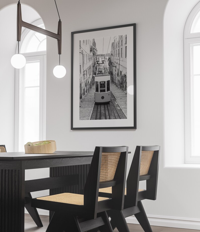 May include: Black and white framed print of a Lisbon tram traveling down a cobblestone street. The image is displayed on a white wall above a black dining table with matching chairs. The chairs have woven cane backs.