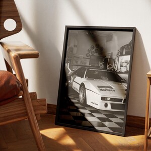 May include: Black and white framed print featuring a vintage race car with the Marlboro logo. The car is parked on a checkered floor. The print is leaning against a wall next to a wooden chair.