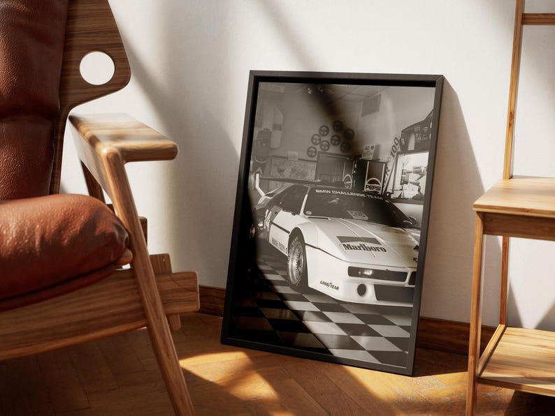 May include: Black and white framed print featuring a vintage race car with the Marlboro logo. The car is parked on a checkered floor. The print is leaning against a wall next to a wooden chair.