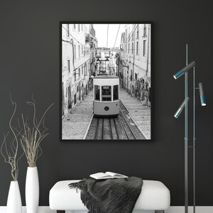 May include: Black and white framed print of a vintage tram car traveling down a cobblestone street. The tram is cream-colored with large windows. The print is displayed above a white bench with a black throw and an open book.