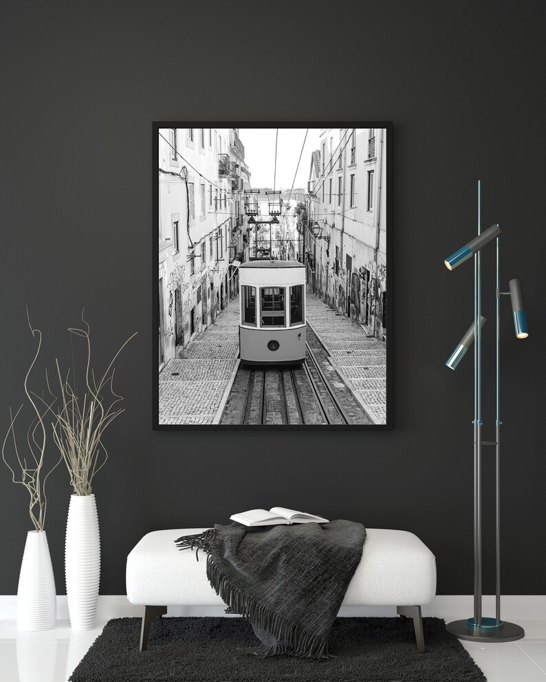 May include: Black and white framed print of a vintage tram car traveling down a cobblestone street. The tram is cream-colored with large windows. The print is displayed above a white bench with a black throw and an open book.