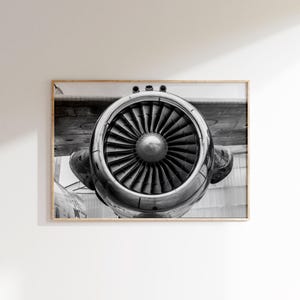 May include: Monochrome photograph of an aircraft engine. The image is framed in a wooden frame. The engine's fan blades are visible, along with the metallic casing and the plane's wing.